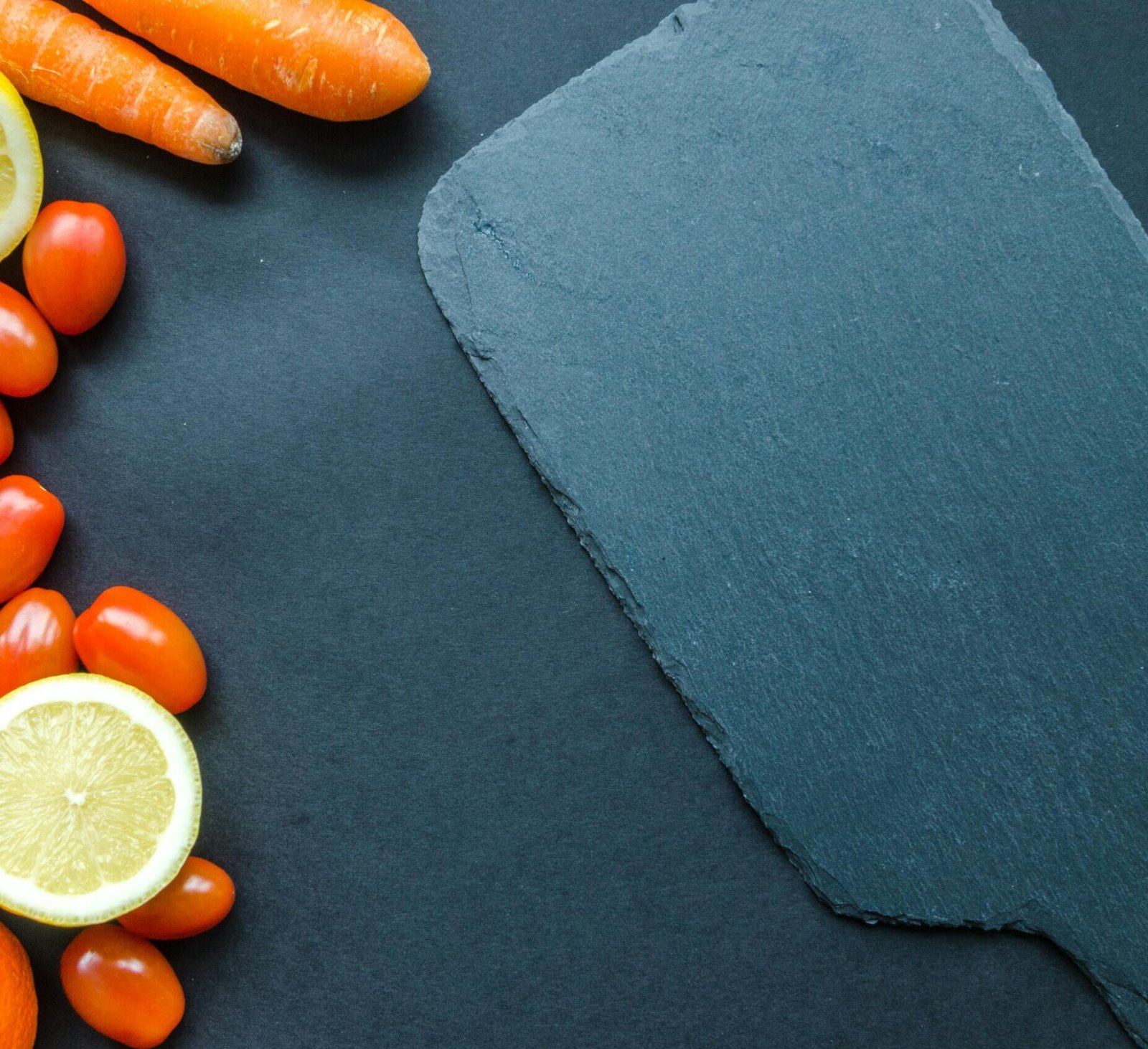 Assortment of fresh fruits and vegetables including carrots, lemon slices, apple, red bell pepper, cherry tomatoes, and an orange on a dark background with space on the right